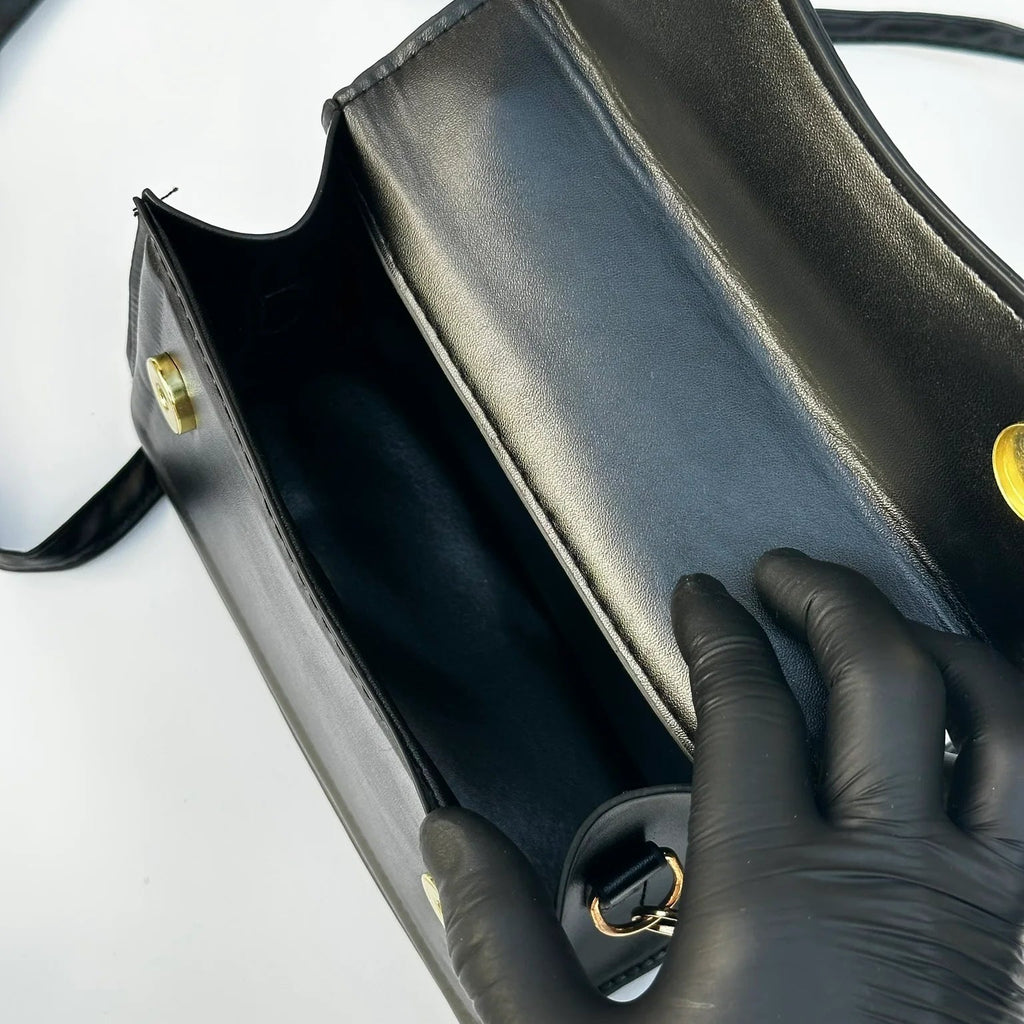 Black handbag with gold hardware being held by a gloved hand on a white background