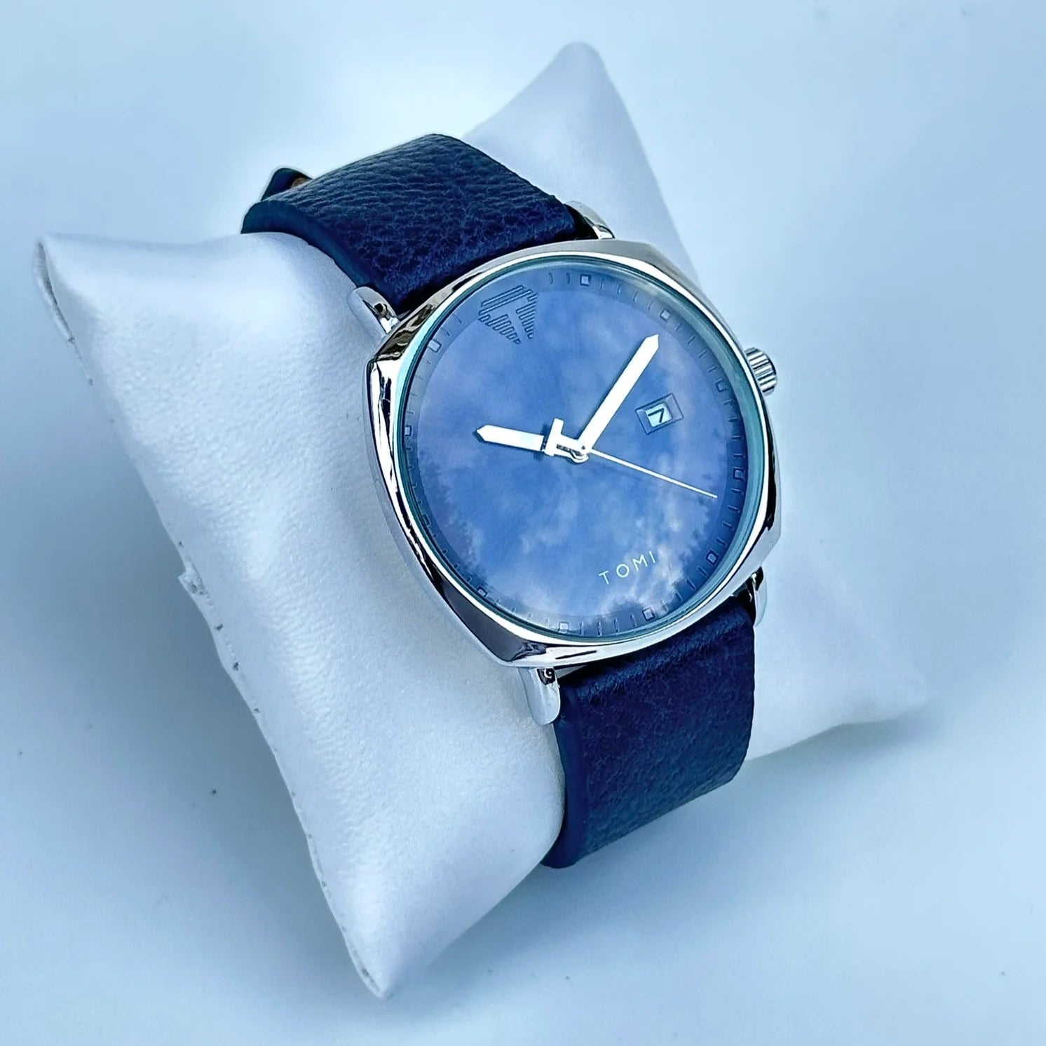 Blue watch with a blue strap on a white stand against a light blue background