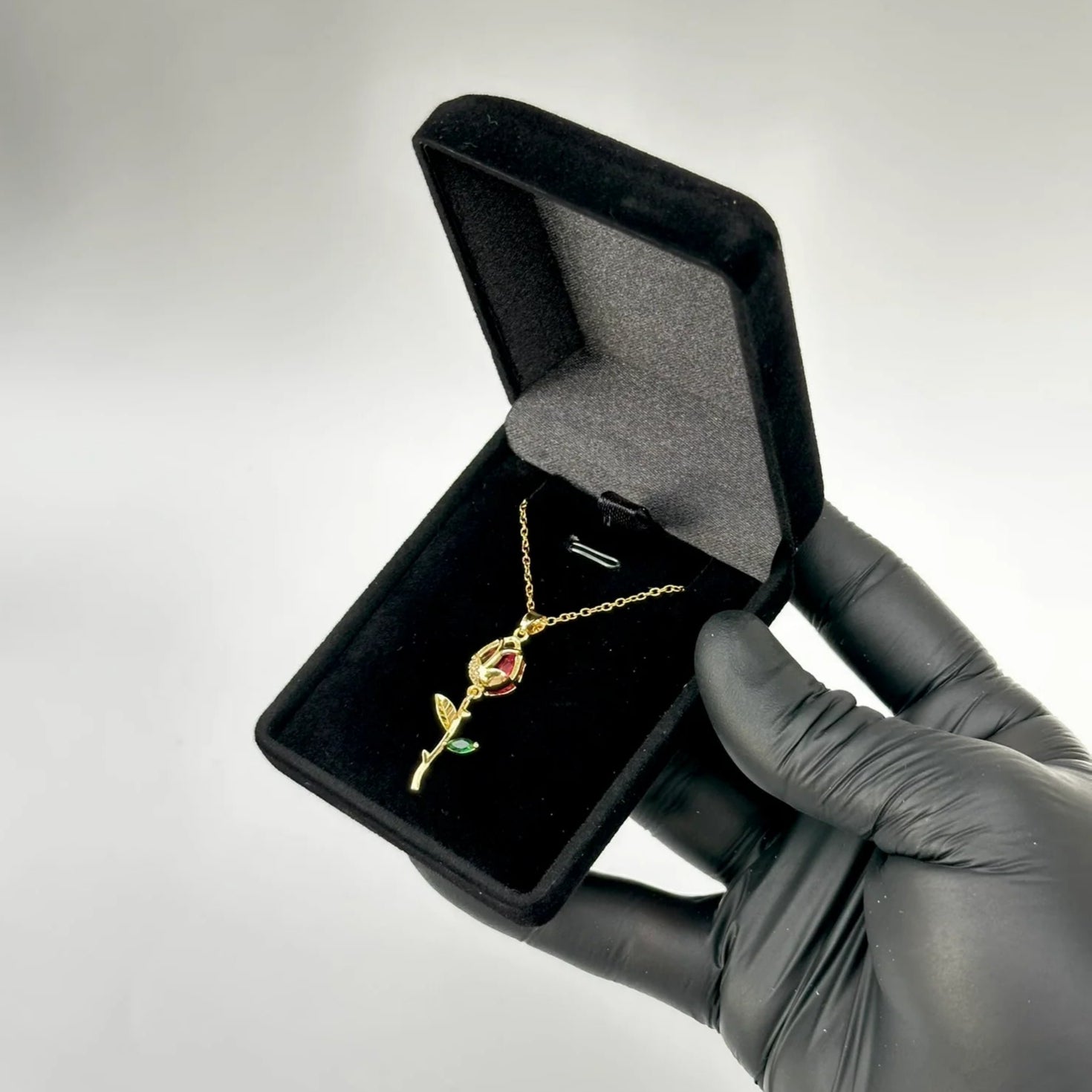 Gold necklace with a red heart pendant in a black jewelry box held by a gloved hand against a gray background