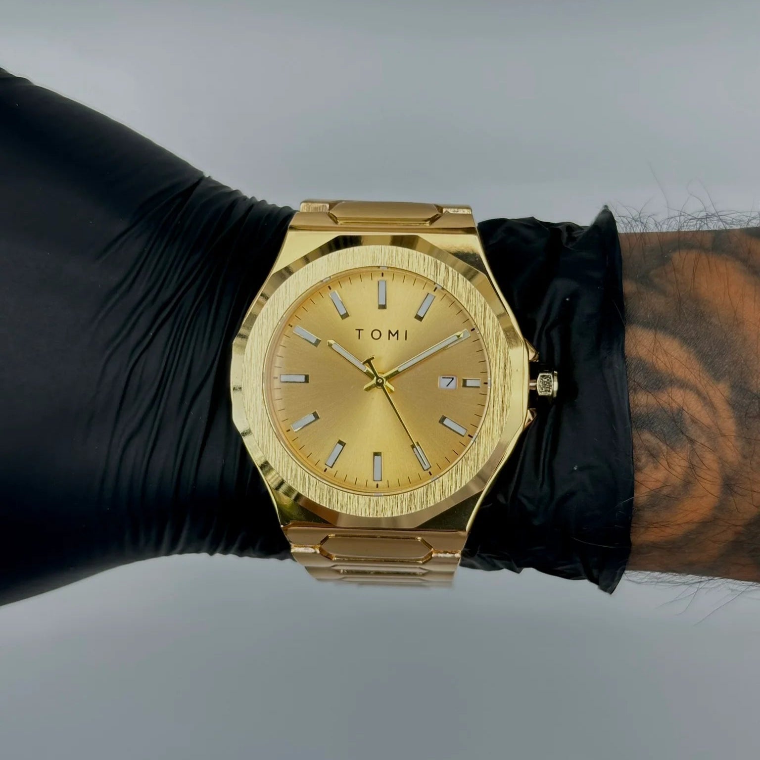 Gold watch on a wrist with a black glove and tattooed arm against a gray background